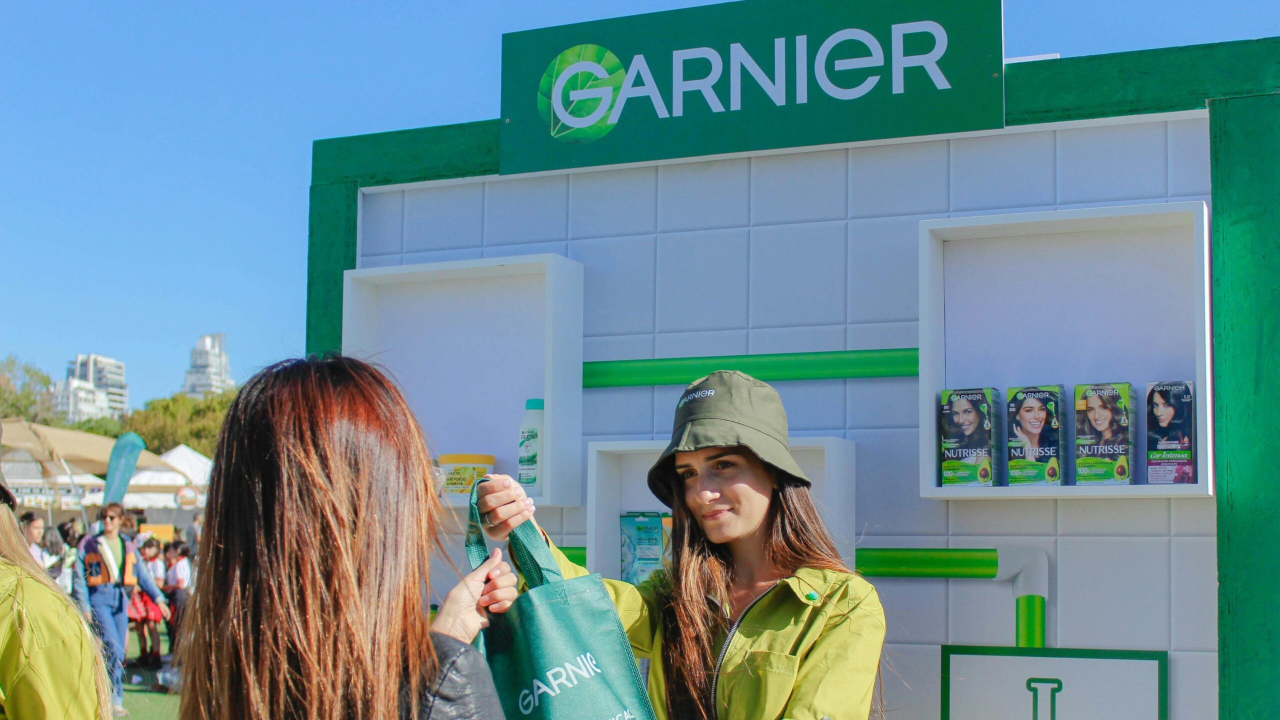 A Garnier brand representative hands a promotional tote bag to an attendee at an outdoor activation booth, with branded products displayed in the background.