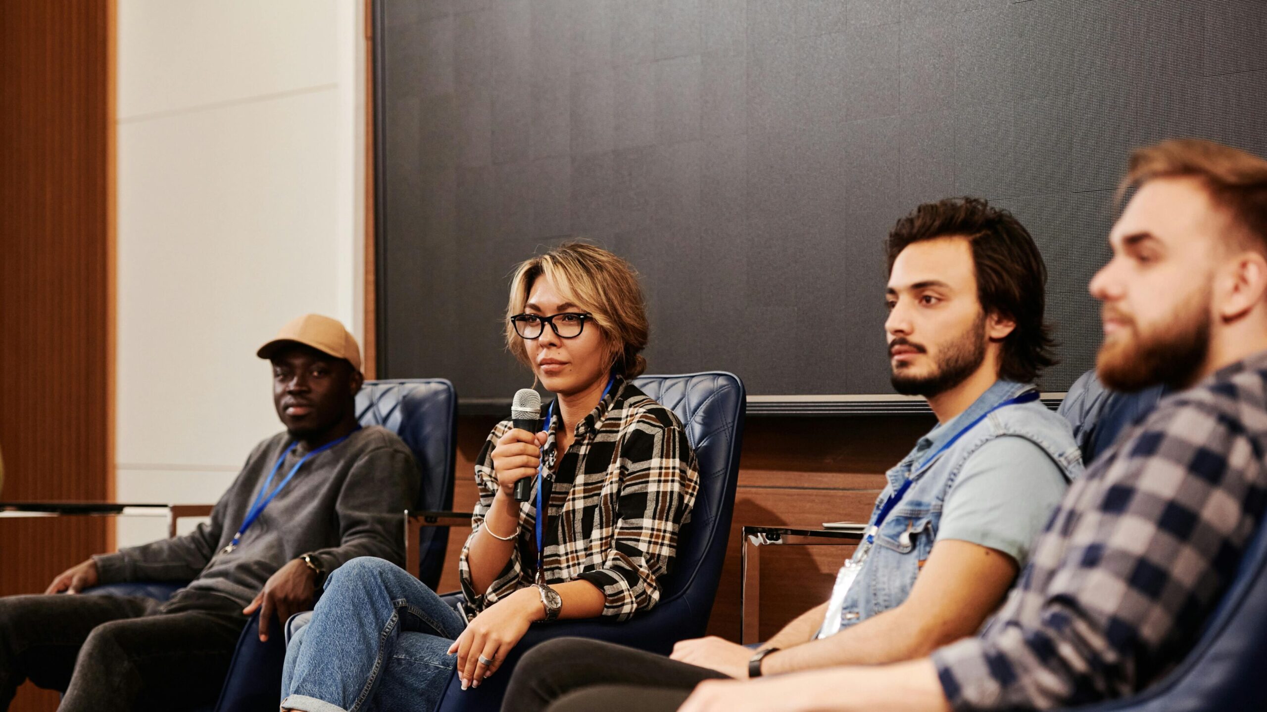 A diverse group of panelists seated in chairs during a conference, with a woman holding a microphone speaking while others listen attentively.