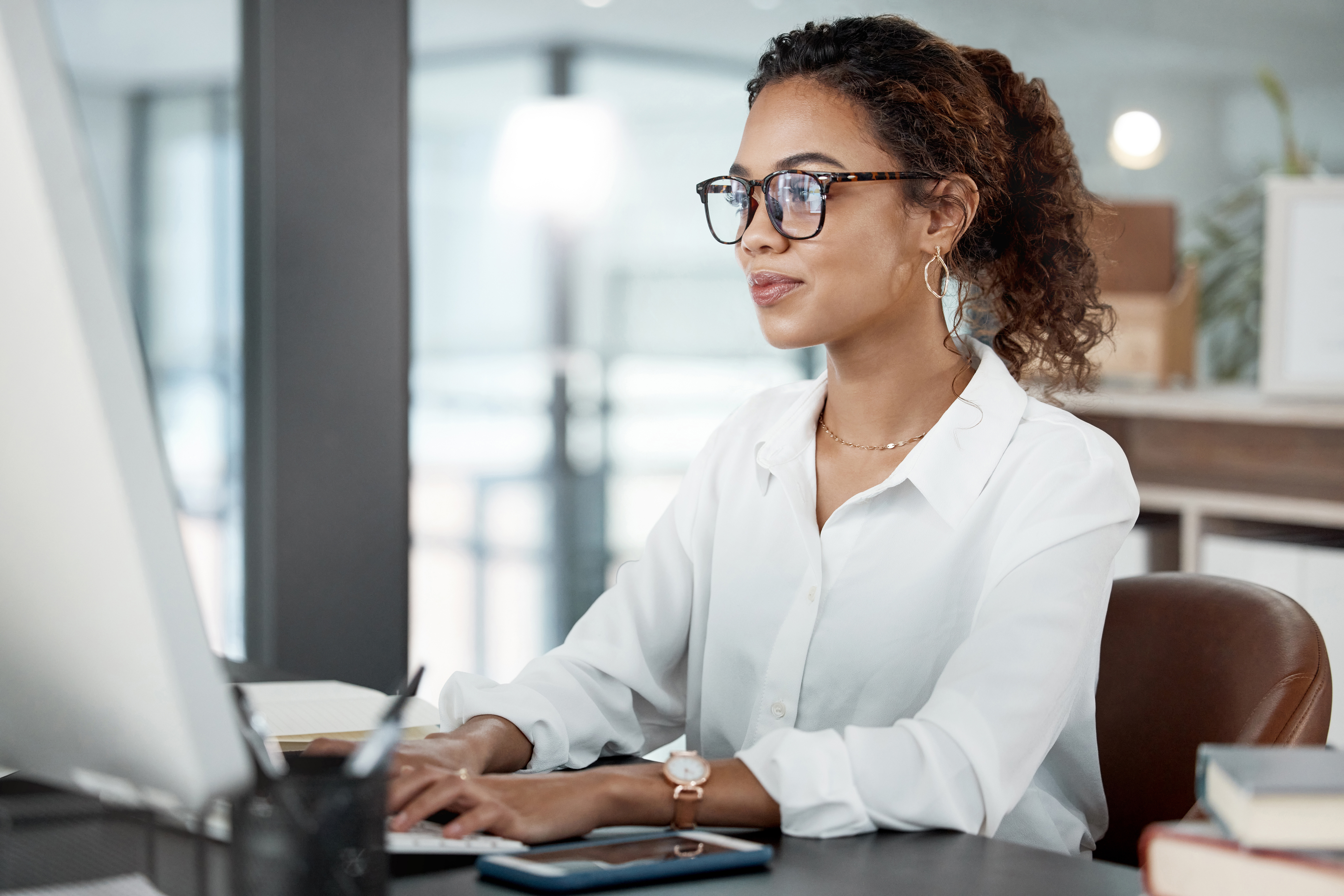 A professional woman wearing glasses and a white blouse sits at a desk working on a desktop computer. She appears focused and confident in a modern office environment, with books, a notebook, and a smartphone on the desk beside her.