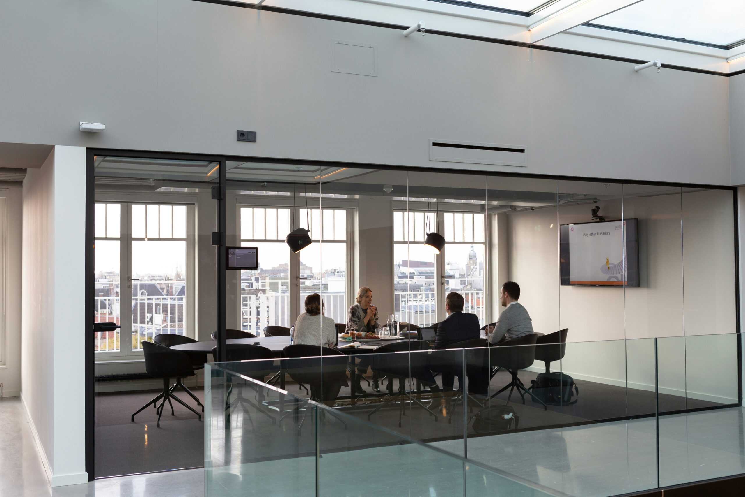 Four people in a modern glass-walled conference room having a business meeting, with a city view visible through large windows.