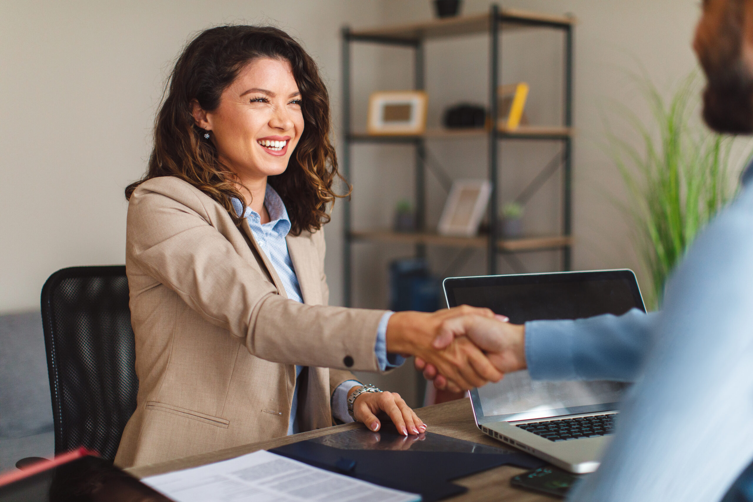 A smiling businesswoman in a beige blazer shakes hands with a colleague across a desk, symbolizing partnership or agreement. The desk has documents, a tablet, and a laptop, and the background features a shelf and a plant, creating a professional office setting.