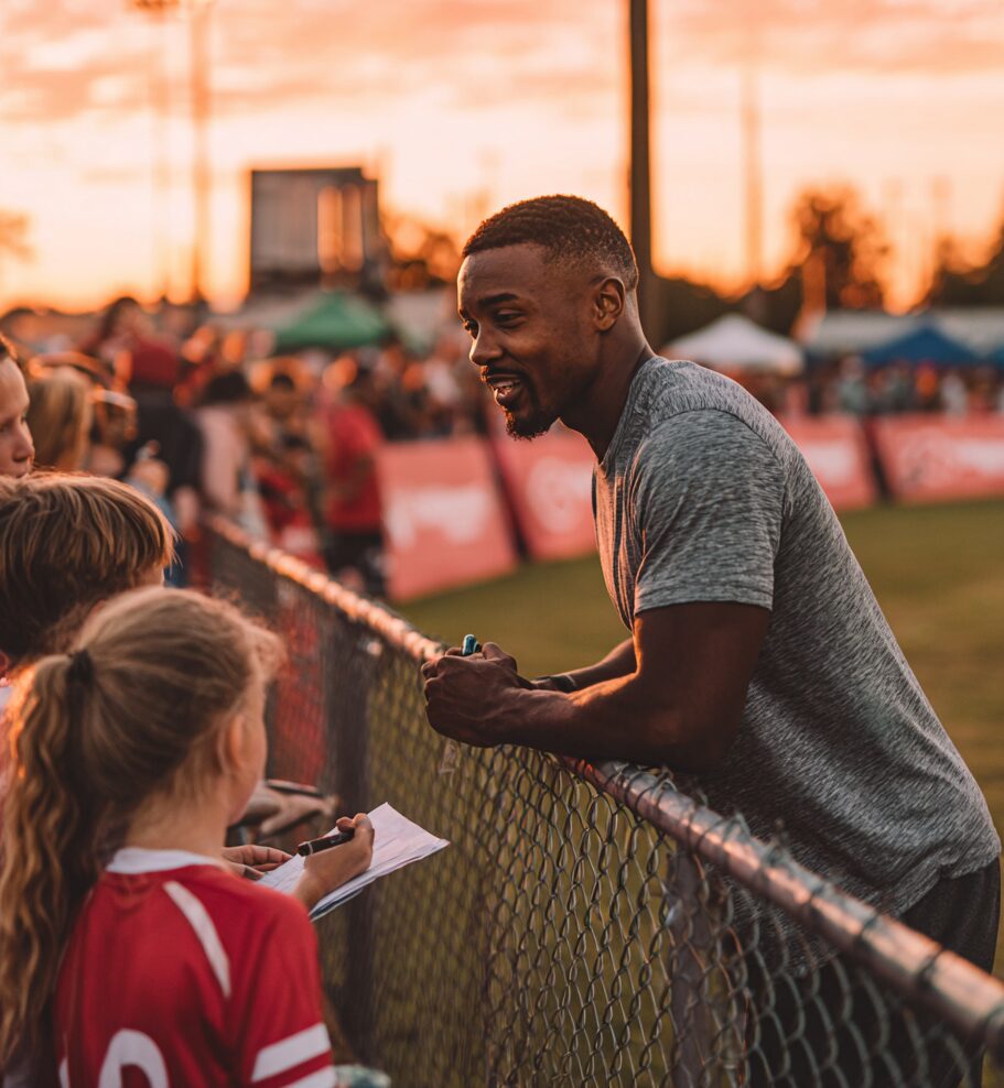 Athlete engaging with young fans at a community sports event during sunset, signing autographs and creating meaningful connections across a fence line.